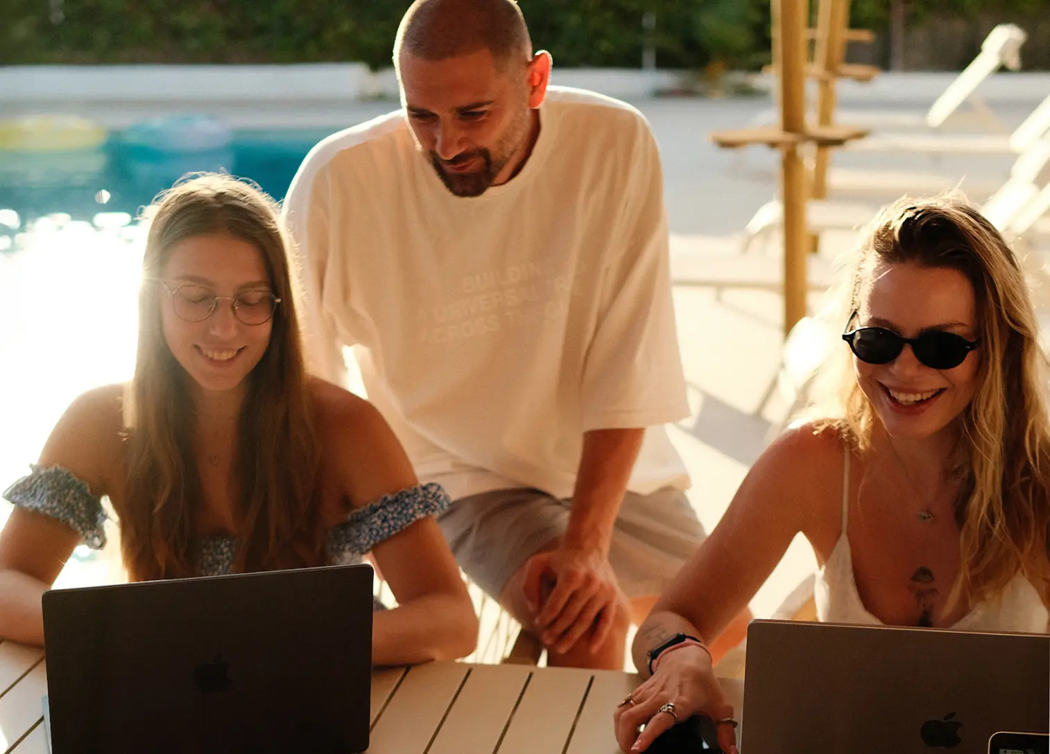 A group of people working on laptops by a pool in a sunny setting.